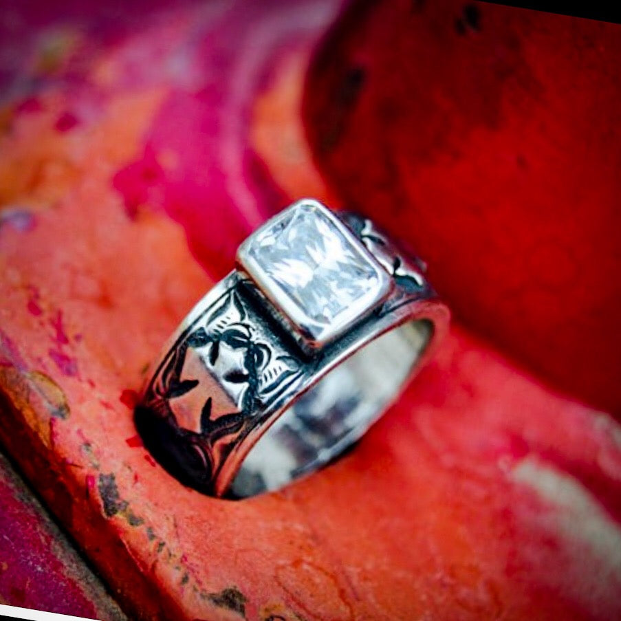 "Lila" is a handmade sterling silver ring that features a bezel set 8mm x 6mm emerald cut cubic zirconia. Its band has western style hand engraving with an overlaid cross on either side of the setting. Because of the width of the band on this ring we recommend ordering it a half size larger than your normal wear.