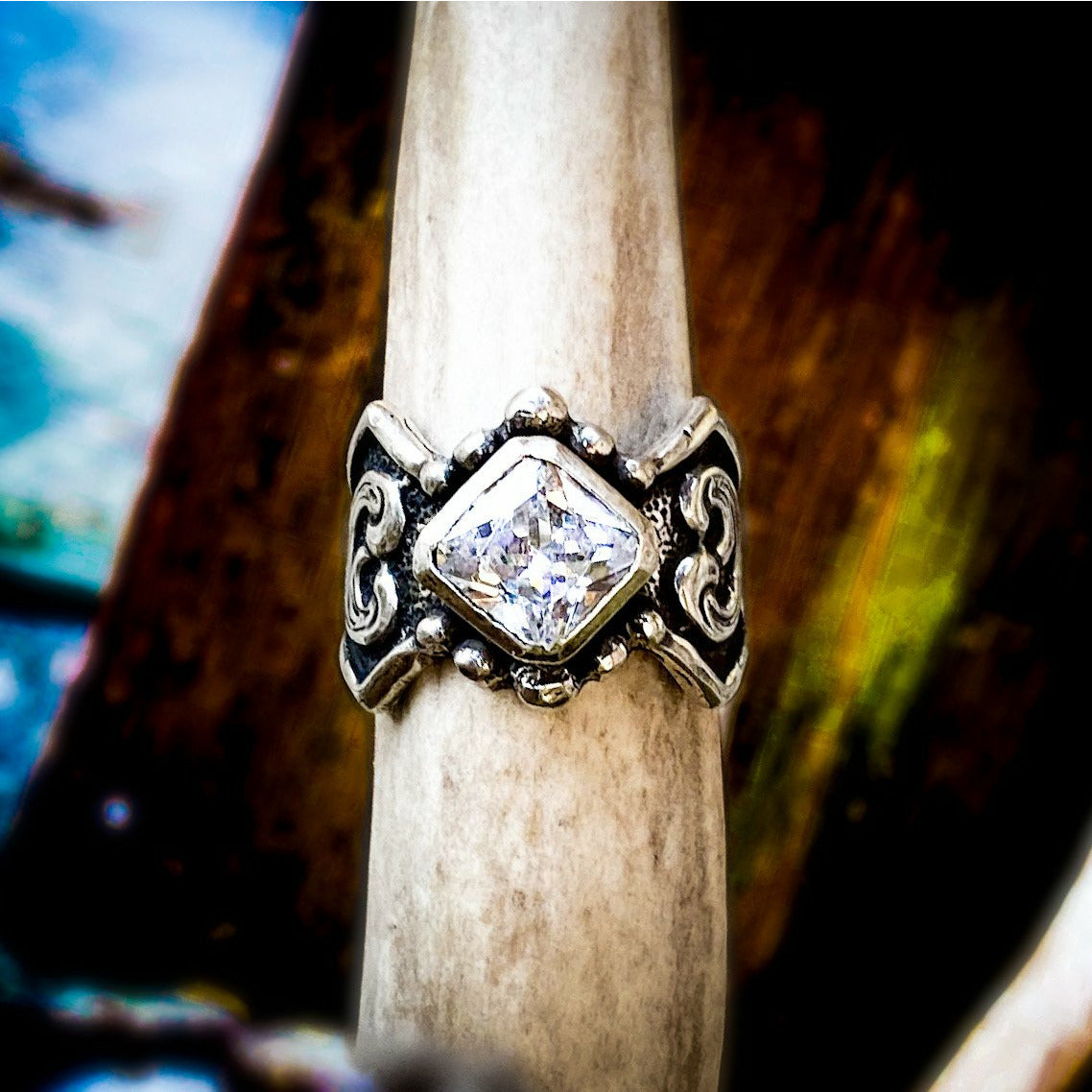 "Jolene" is a custom sterling silver ring that has a unique border surrounding an 8mm bezel set asher cut cubic zirconia mounted in the center of the ring and features a western style engraved scrolling overlay on either side of the band. 
