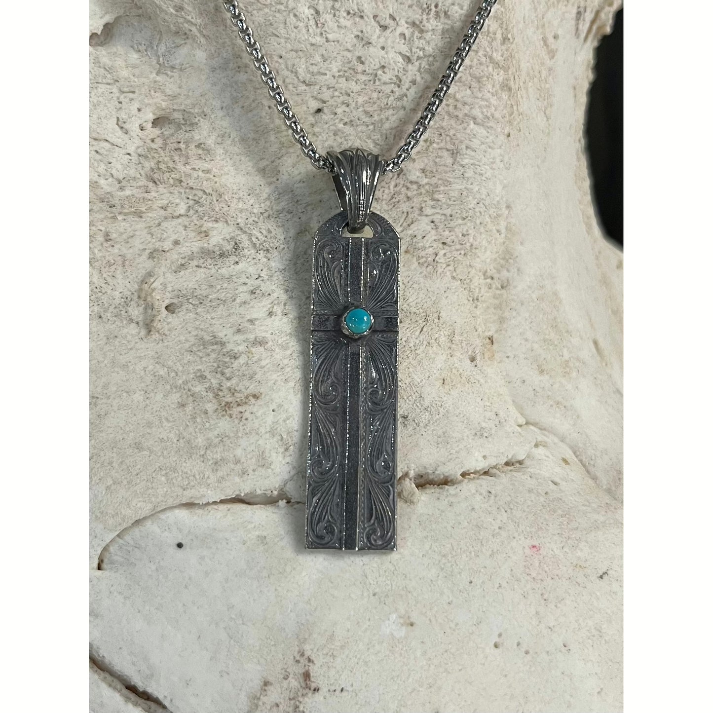 The "Grace" Pendant is a handmade sterling silver pendant with a western style hand engraved scrolling with an overlaid cross that features a bezel set round Kingman turquoise stone from the Kingman Mines in Kingman, AZ. It measures 2 7/8" long and 1/2" wide.