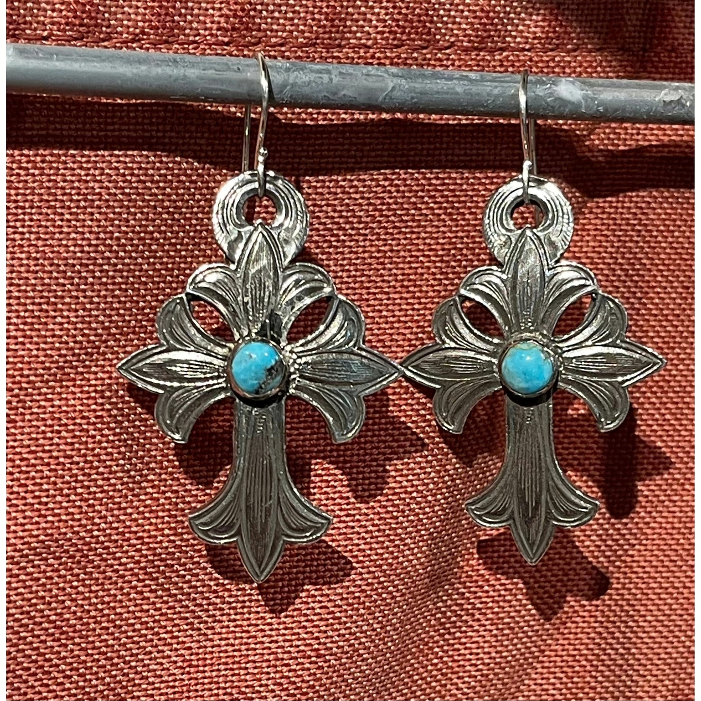 The "Mary" Cross Turquoise Earrings are handmade solid sterling silver dangle earrings that feature western style hand engraving and set with a genuine Kingman stone in the center of the cross. These earrings are 1 1/4" X 1 3/4".