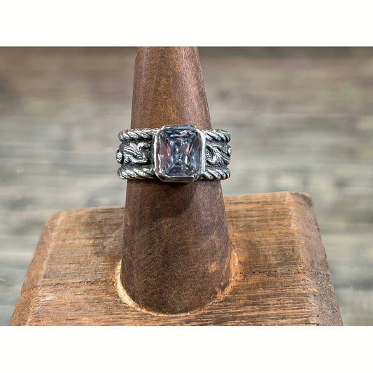 "Morgan" is a handmade sterling silver ring that features a 9mm x 7mm emerald cut cubic zirconia in a heavy bezel setting. The unique band has a thick roped edge all the way around the band that surrounds its beautiful overlaid scrolling on either side of the band.