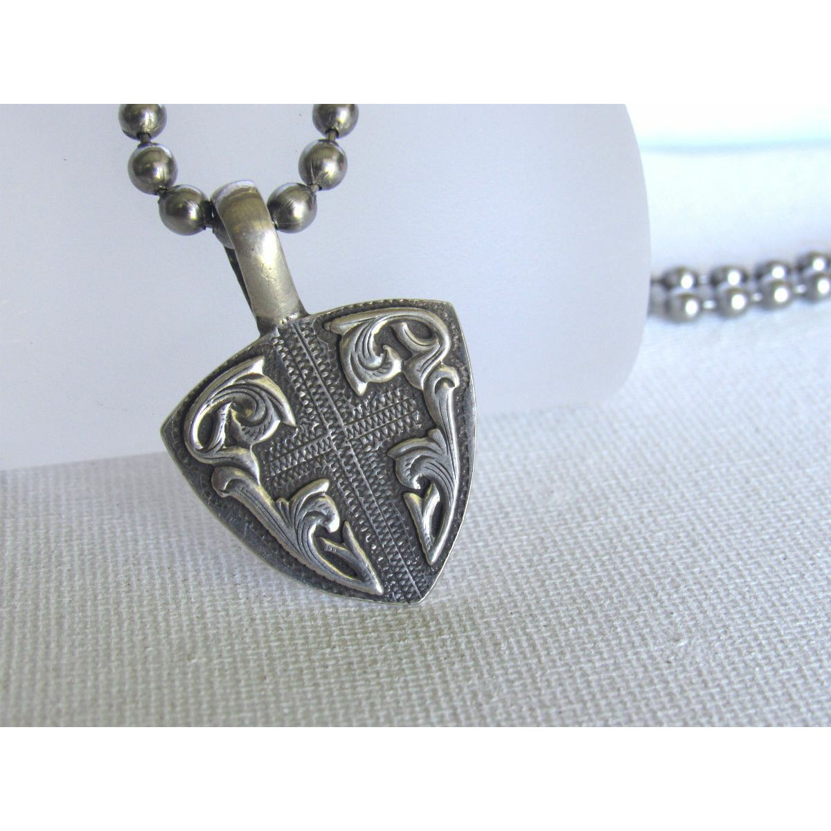 The "Hidden Cross Guitar Pick" is a handmade solid sterling silver guitar shaped pendant with a smooth raised border with an edgy western style hand engraved background which features a smooth recessed cross. The pendant measures at 1" X 1 3/8".
